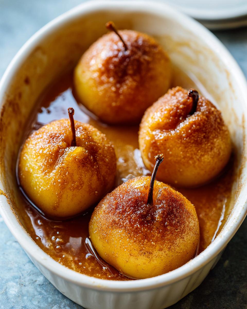 Close-up of four glistening baked apples dusted with cinnamon sugar, resting in a syrupy sauce in a white baking dish.