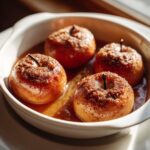 Four perfectly baked apples topped with cinnamon sugar, sitting in a sweet syrup in a white dish.