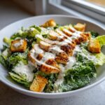 A close-up of a delicious caesar salad topped with sliced grilled chicken, croutons, and creamy dressing.