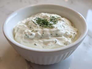 A close-up of creamy homemade tartar sauce garnished with fresh dill in a white ribbed bowl.