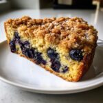 A close-up slice of moist blueberry breakfast cake featuring a thick, golden crumb topping dusted with powdered sugar.