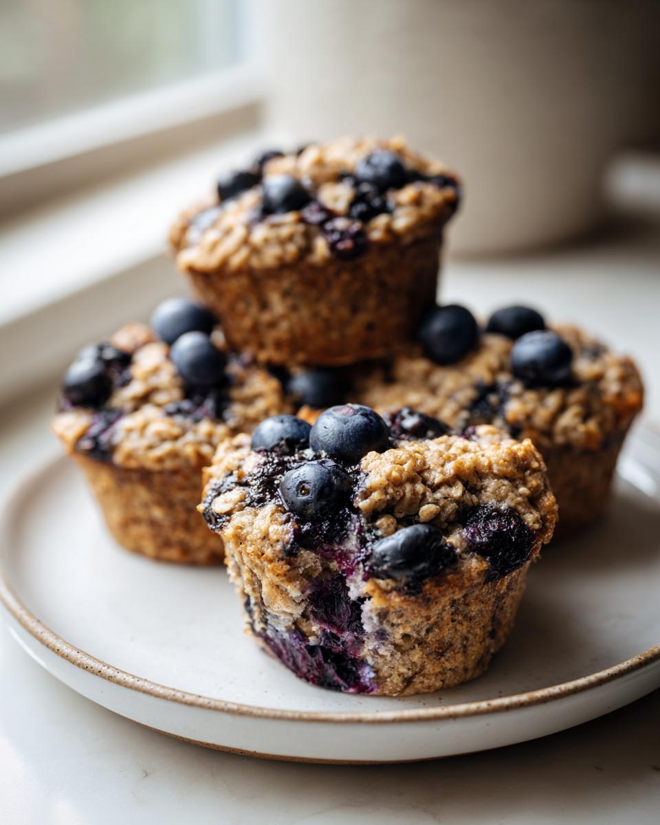 A close-up of several moist blueberry oatmeal muffins topped with fresh blueberries, served on a light plate.