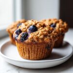 A close-up of a perfectly baked blueberry oatmeal muffin with a crumbly top, sitting on a white plate.