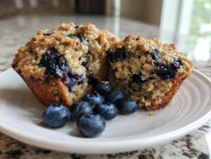 A close-up of a blueberry oatmeal muffin cut in half, showing the texture and fresh blueberries inside, served with whole blueberries.