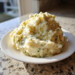 A generous serving of creamy, fluffy boursin mashed potatoes flecked with green herbs on a white plate.