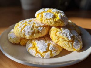 A stack of bright yellow lemon crinkle cookies dusted heavily with white powdered sugar, resting on a white plate.