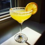 A bright yellow lemon drop cocktail in a coupe glass with a sugar rim and lemon slice garnish.