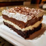 A rich, layered slice of brownie refrigerator cake featuring a dark brownie base, creamy white layer, chocolate mousse, and whipped topping.