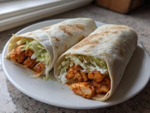 Two tightly rolled buffalo chicken wrap halves filled with spicy chicken, shredded lettuce, and white sauce, served on a white plate.