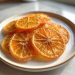 A close-up shot showing several glistening candied orange slices dusted with sugar resting on a speckled white plate.