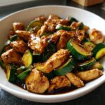 A close-up of delicious chicken zucchini stir fry coated in a dark, glossy sauce and sprinkled with sesame seeds.