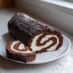 A chocolate yule log cake, textured to look like bark, sliced to show the white cream swirl filling.