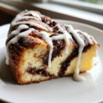 Close-up of a moist slice of cinnamon roll cheesecake with dark cinnamon swirl and thick white icing drizzled over the top.