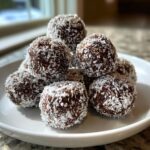 A close-up stack of rich, dark bourbon balls heavily coated in shredded white coconut flakes.