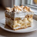 A tall, square slice of creamy banana pudding layered with vanilla wafers and fresh bananas, topped with toasted meringue.