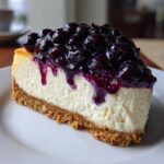 A perfect slice of creamy blueberry cheesecake sits on a white plate, generously topped with rich blueberry compote.