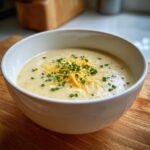 A close-up of a white bowl filled with creamy cauliflower soup, garnished with shredded cheddar cheese and fresh chopped chives.