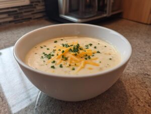 A close-up of a white bowl filled with creamy cauliflower soup, topped with shredded cheddar cheese and fresh chopped chives.
