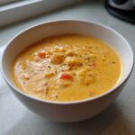 A close-up of a white bowl filled with rich, orange cheese soup containing chunks of ingredients and topped with dried herbs.