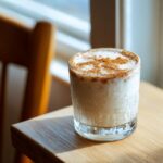 A glass of creamy, white coquito topped generously with ground cinnamon, sitting on a wooden table near a window.