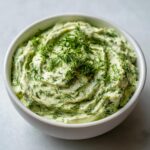 A close-up of a creamy, light green dill dip generously topped with fresh, feathery dill in a white bowl.