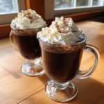 Two glass mugs filled with rich, dark hot chocolate topped generously with whipped cream and a dusting of cinnamon.