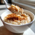 A spoonful of creamy rice pudding recipe topped with cinnamon being lifted from a white bowl.