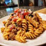 A close-up of creamy taco pasta salad featuring rotini pasta, seasoned ground beef, and topped with fresh tomatoes, red onion, and black olives.