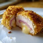 Close-up of a perfectly cooked chicken cordon bleu cut in half, showing melted cheese oozing out over ham inside the crispy breading.