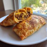 Two golden-brown, crispy Chimichangas on a white plate, one cut open to show ground beef and melted cheddar cheese filling.