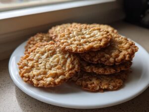 A stack of golden brown, crispy oatmeal lace cookies resting on a small white plate near a window.