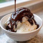 A scoop of vanilla ice cream in a white bowl being generously drizzled with thick, glossy chocolate syrup.