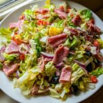 A vibrant close-up of a fresh grinder salad featuring chopped lettuce, rolled deli meats, red onion, black olives, and dressing.