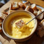 A piece of crusty bread being dipped into a bowl of smooth, yellow cheese fondue recipe using a fondue fork.