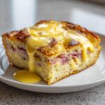 A thick slice of baked eggs benedict casserole layered with ham, topped generously with dripping hollandaise sauce.