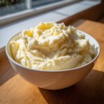 A large white bowl filled with incredibly fluffy mashed potatoes, highlighted by sunlight.