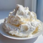 A large, fluffy mound of bright white vanilla buttercream frosting sitting on a white plate, catching bright sunlight.