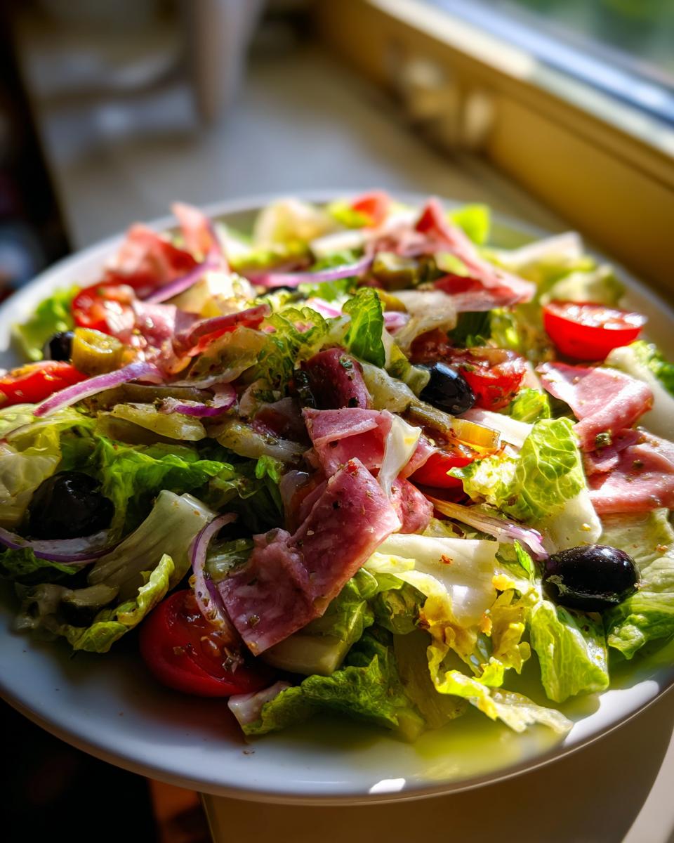 Close-up of a vibrant grinder salad featuring lettuce, deli meats, tomatoes, olives, and red onion, brightly lit.