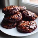 A stack of freshly baked, fudgy chocolate brownie cookies featuring glossy, cracked tops, resting on a white plate.