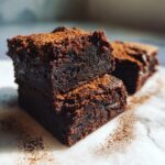 Close-up of two fudgy date brownies stacked, dusted with cocoa powder on a light surface.