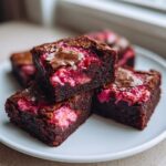 Close-up of fudgy strawberry brownies stacked on a white plate, showing bright pink strawberry swirls inside.