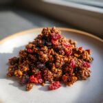 A mound of dark, rich gingerbread granola mixed with dried cranberries served on a light gray plate.