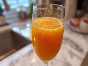 Close-up of a sparkling, orange bellini recipe cocktail in a champagne flute on a kitchen counter.