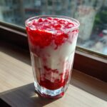 A tall glass showing layers of white milk and bright red strawberry puree, making delicious strawberry milk.