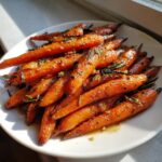 A plate piled high with glossy, caramelized roasted carrots seasoned with fresh rosemary.