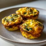 Three golden brown, baked egg bites with visible spinach pieces resting on a speckled ceramic plate.