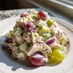 A mound of creamy greek yogurt chicken salad mixed with celery, red onion, and whole red and green grapes on a white plate.
