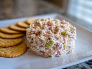 A molded serving of creamy ham salad recipe with visible celery and ham pieces, served alongside round crackers on a white plate.