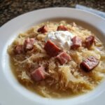 A close-up of a white bowl filled with hearty sauerkraut soup, topped with chunks of smoked sausage and a dollop of sour cream.