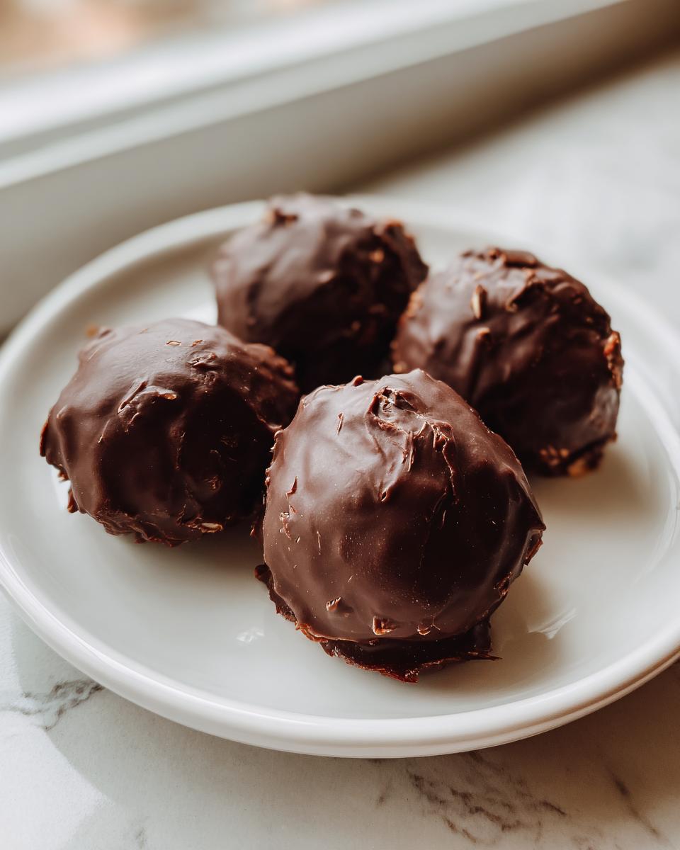 Four rich, chocolate-covered almond joy truffles resting on a white plate near a window.